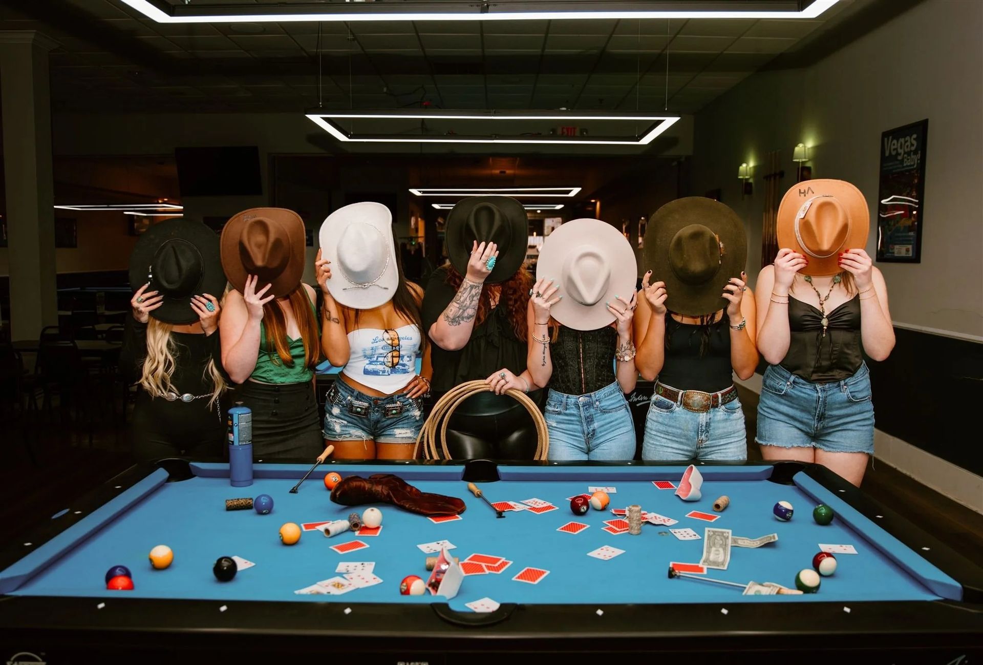Six masked people stand behind a pool table covered with snacks and drink cans in a dim room