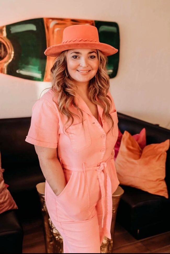 Woman in a coral outfit and hat standing indoors beside a black couch with orange pillows.