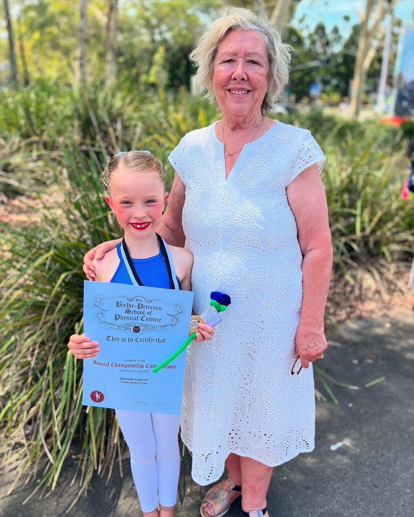 Girl in blue leotard with award, smiles with teacher in white dress. Outdoors, sunny day.