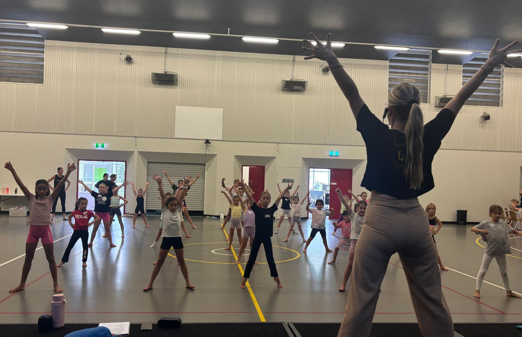 Dance class with children and instructor in a large gymnasium; arms raised.