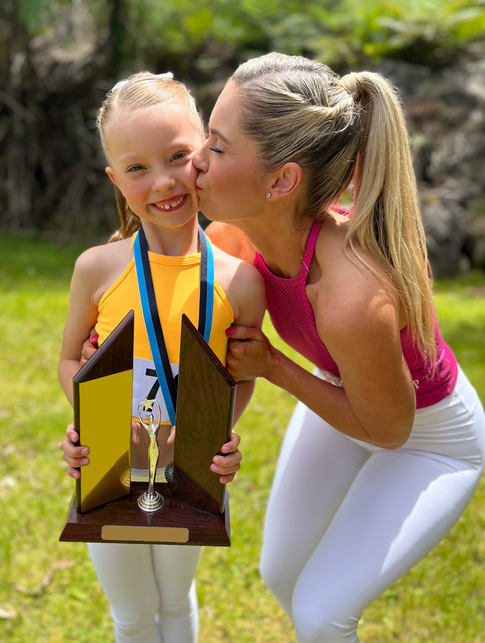 A young girl, holding a trophy, gets kissed on the cheek by her mum. Both are smiling, outdoors.