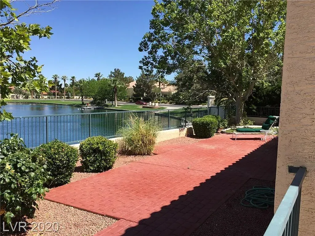 Red gravel patio beside a calm lake, with green shrubs, trees, and a blue sky.