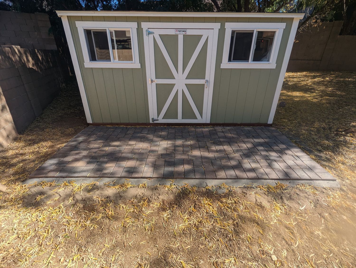 A green shed with white trim and windows is sitting on a brick patio.