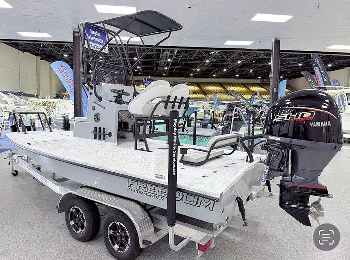 New 2026 Freedom Commander shallow water fishing boat on a trailer at an indoor boat show, with a T-top and outboard motor
