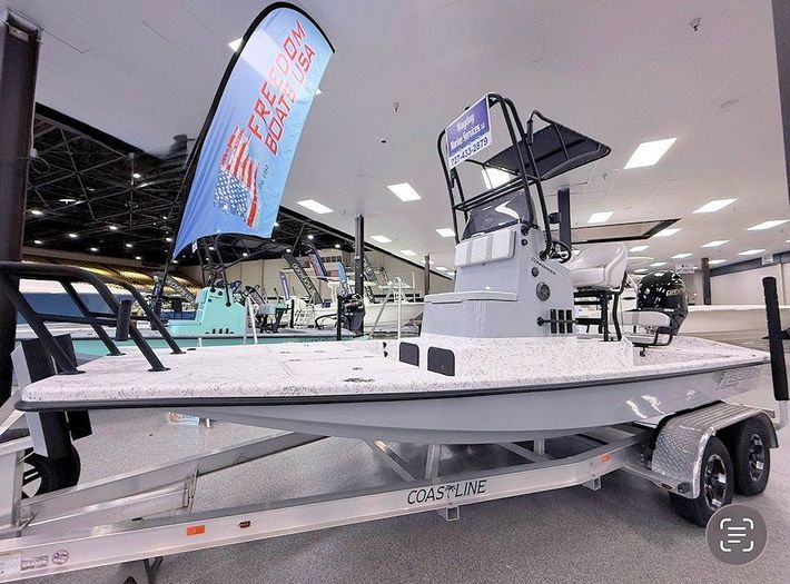 New 2026 Freedom Commander shallow water fishing boat on display indoors with a blue flag and white hull covered in sponsor logos