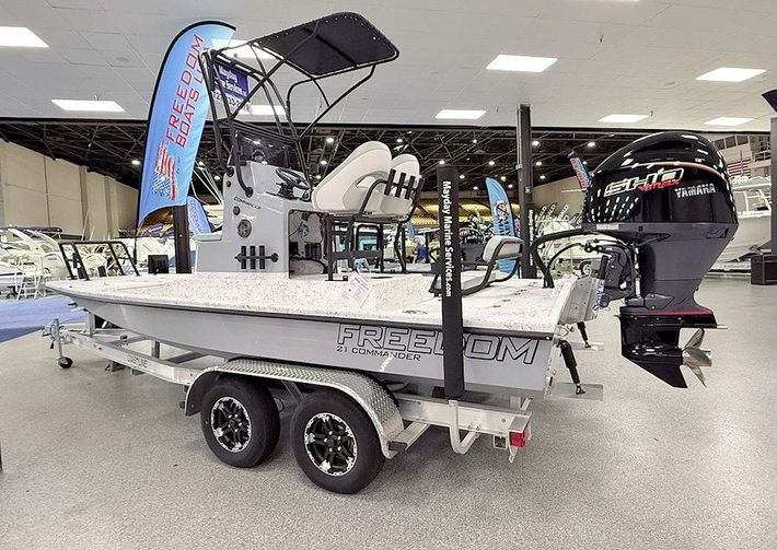 New 2026 Freedom Commander shallow water fishing boat on a trailer in a showroom, with a black outboard motor and fishing gear.
