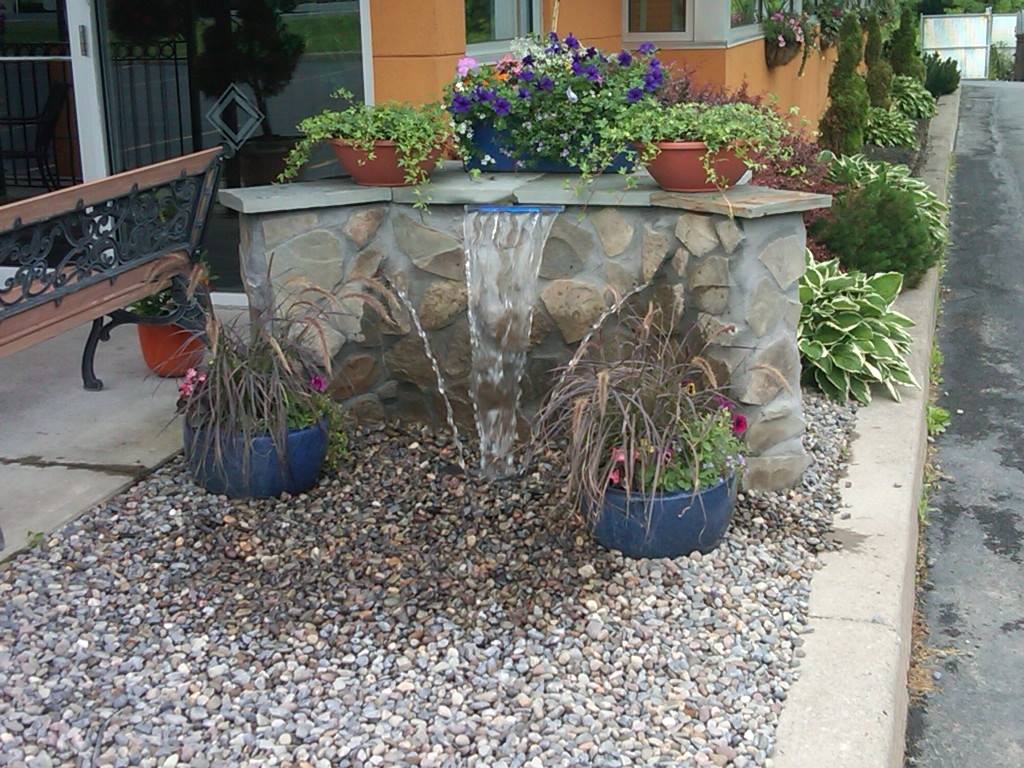 Water fountain with stone facade, surrounded by planters with flowers and gravel.