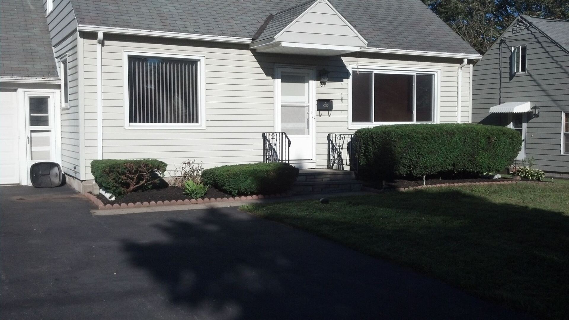 Cottage-style house with light siding, dark roof. Front door centered between two windows. Small manicured bushes in front.