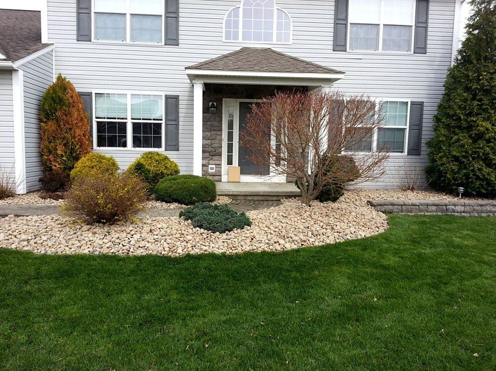 Front of a two-story house with landscaping. Green grass in front, rock bed with shrubs.