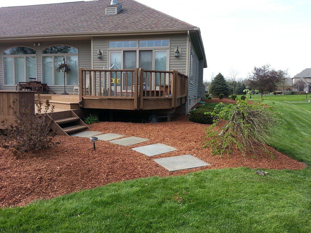 Wooden deck and stone pathway leading to a house with a grass lawn. Mulch and plants in the foreground.