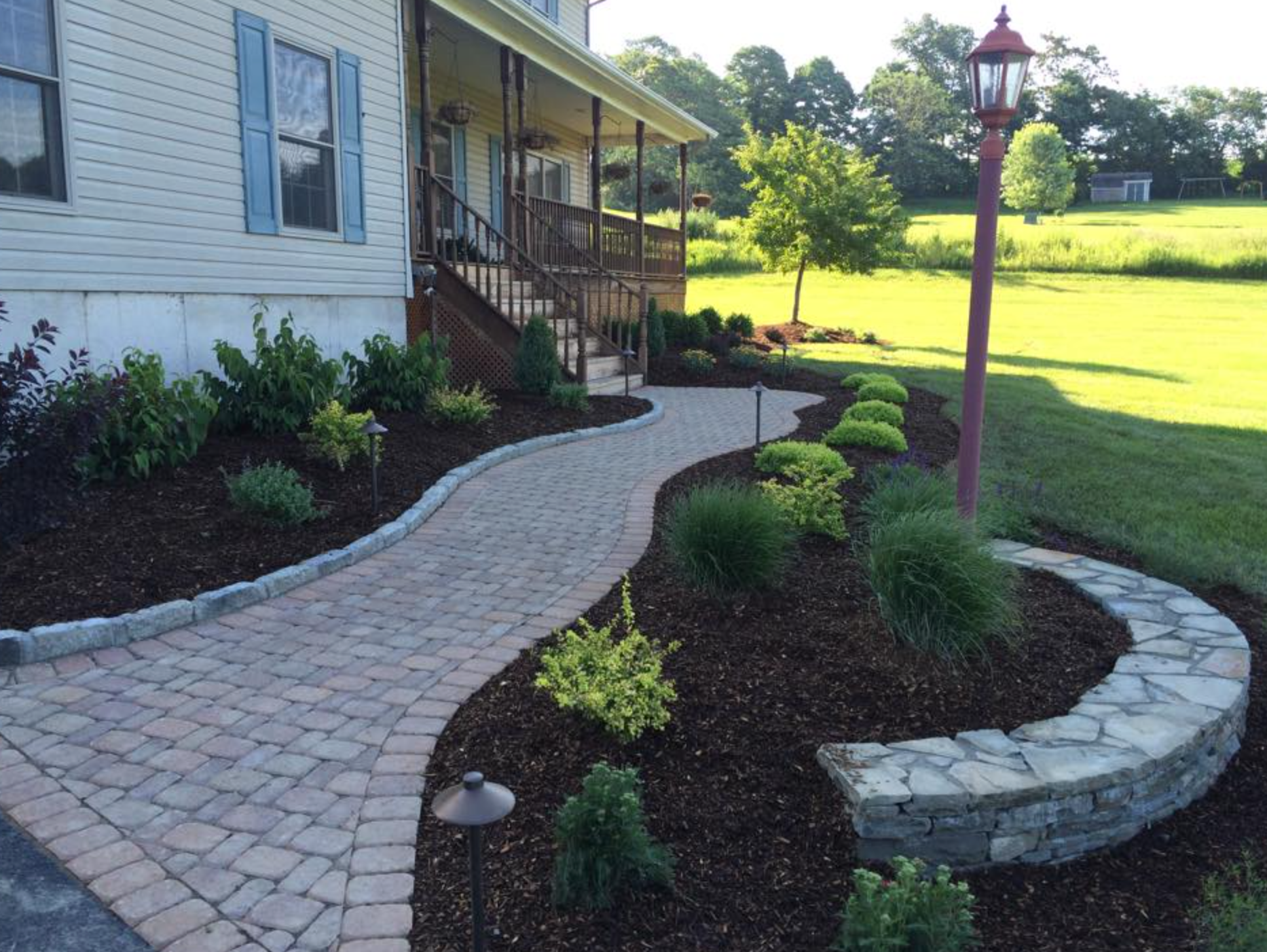 Brick pathway curving to a house with a porch, bordered by landscaped beds, and a decorative stone wall.