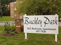 Sign for Buckley Park apartments, with green lettering, white background, and phone number, with apartments in the background.