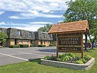 Hampton Estates apartment complex sign on green grass, brown building. Cloudy blue sky.
