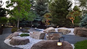 Zen garden with large rocks, white gravel, trees, and small lanterns.