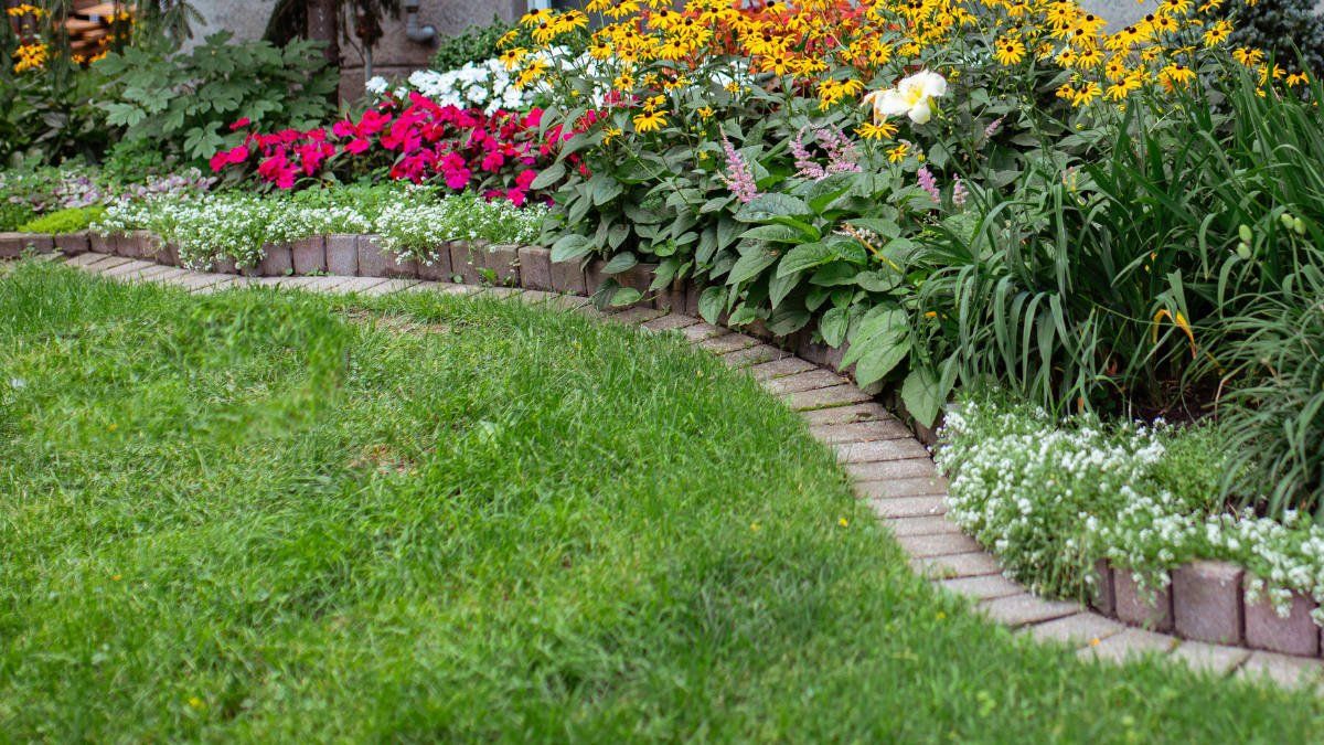 Lush green grass curving alongside a flowerbed filled with various colorful blooms, bordered by a brick pathway.