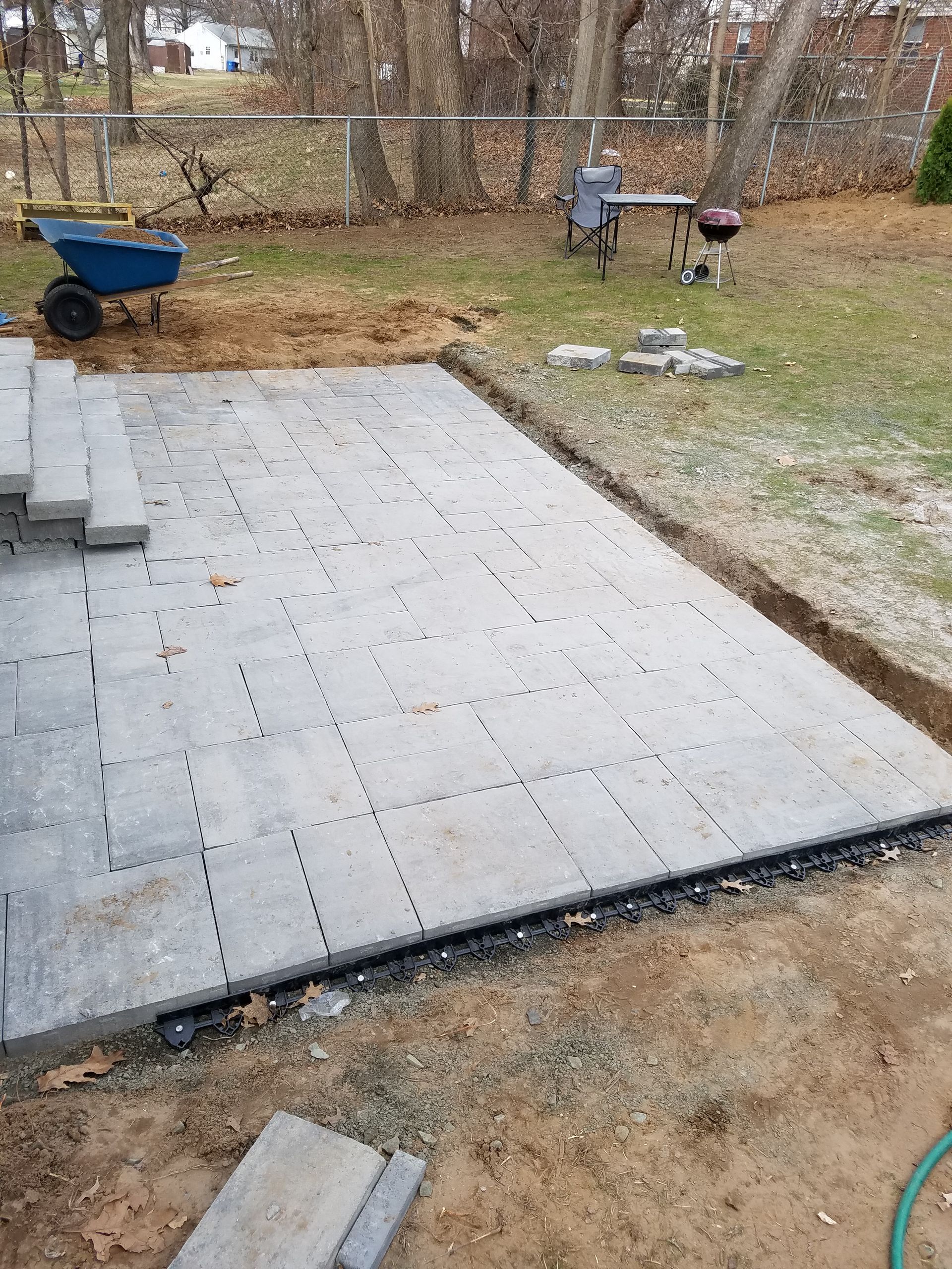 A newly installed rectangular gray paver patio in a backyard, with a wheelbarrow nearby and a trench along the edge.