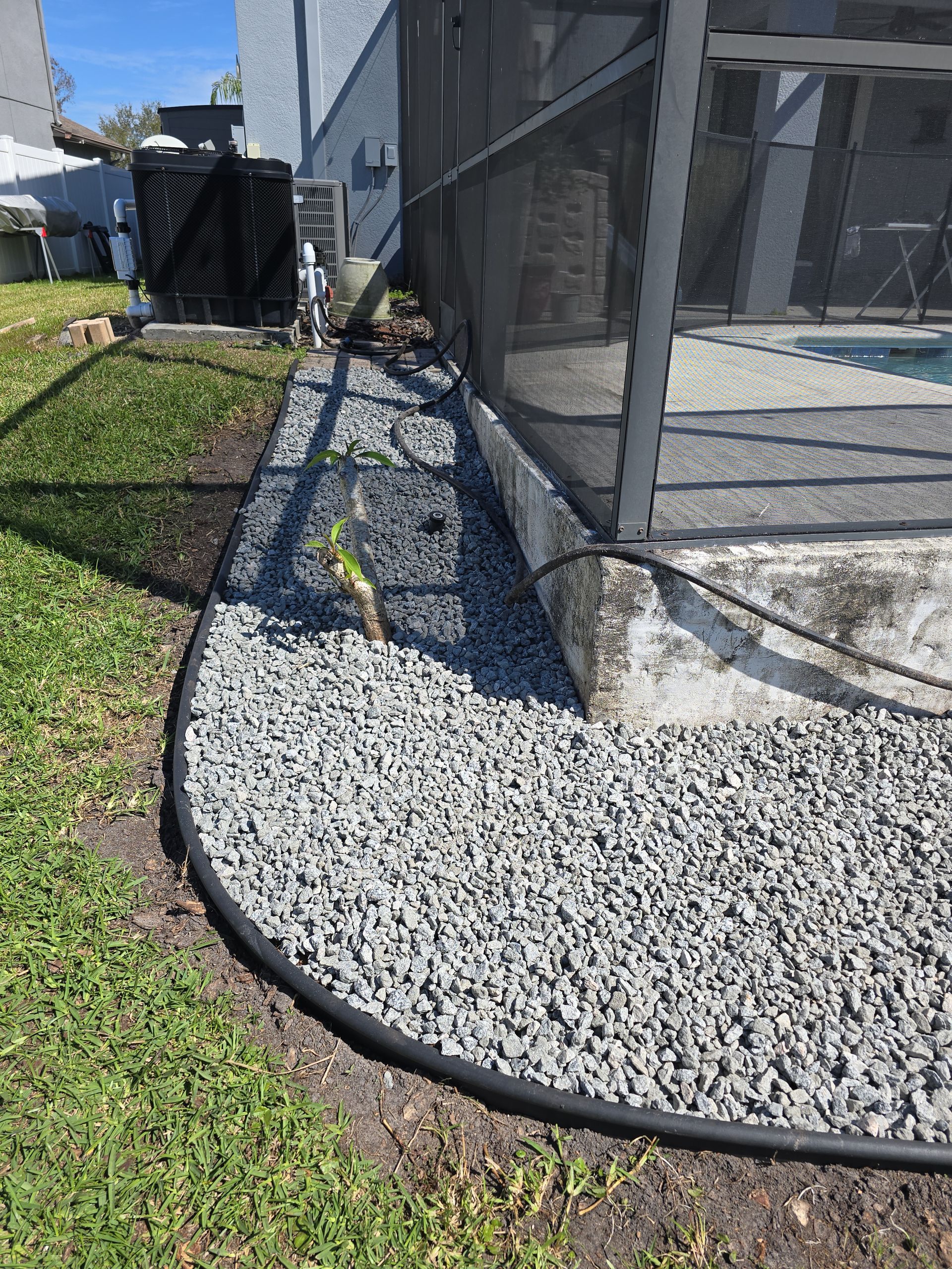 A curved landscape border filled with gray gravel next to a pool screen enclosure and a green lawn.