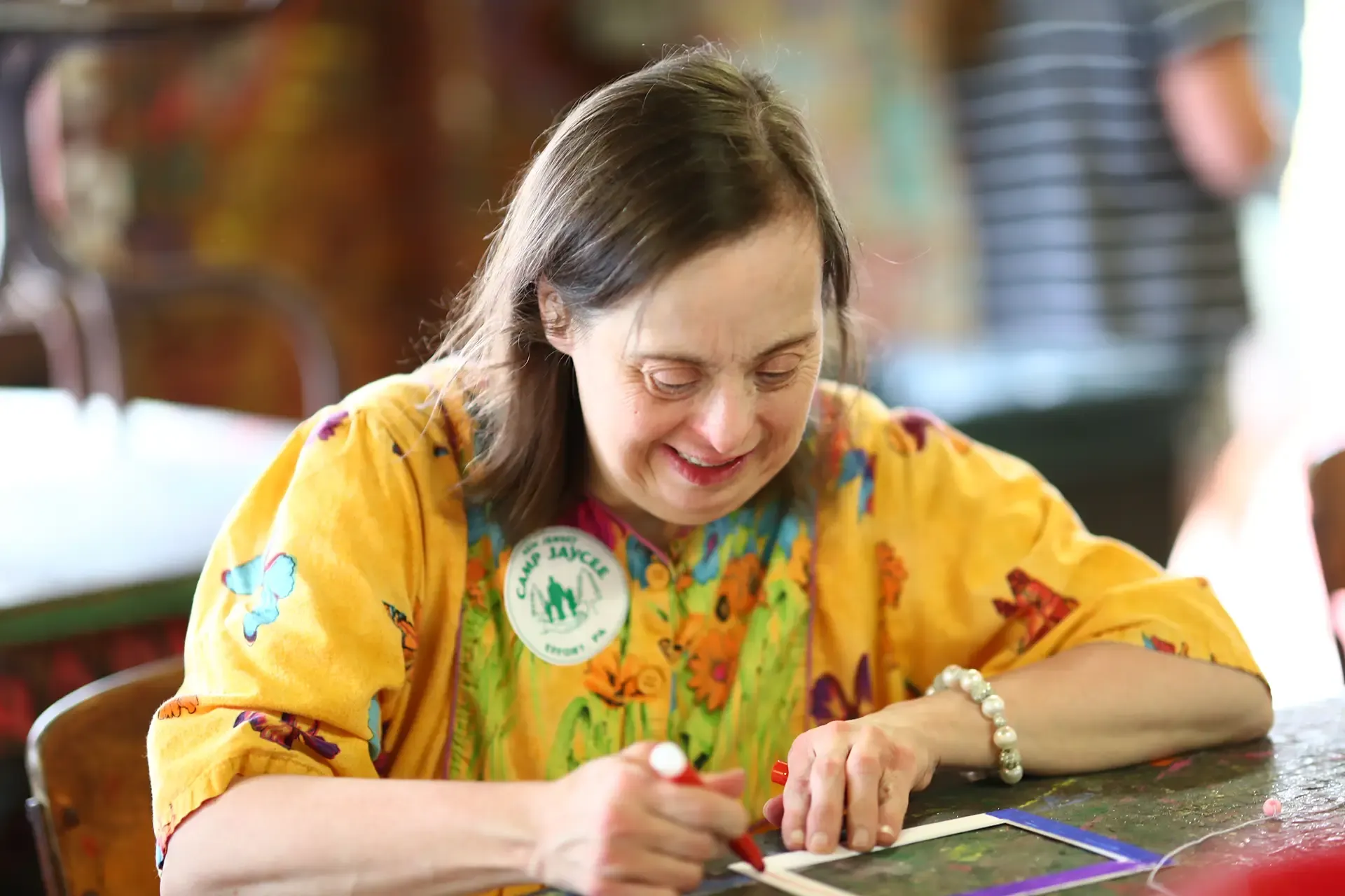 Woman with Down syndrome signing a paper, wearing a yellow floral shirt, indoors.