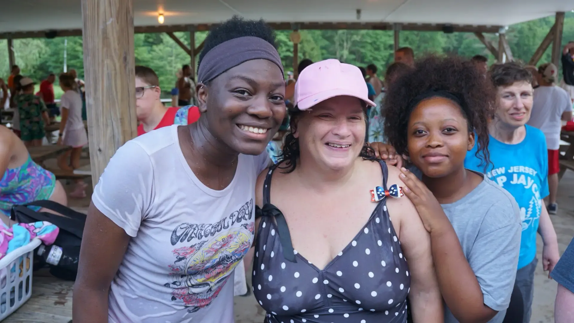 Three people smiling, one wearing a pink hat, another a headband, outside under a covered area.