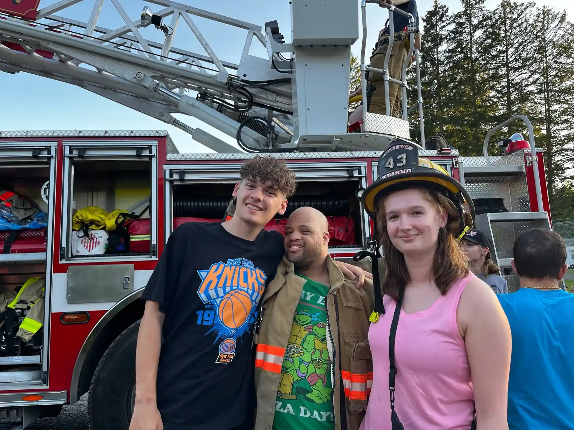 Three people pose with a fire truck. Two are smiling. A firefighter wears a helmet and coat.