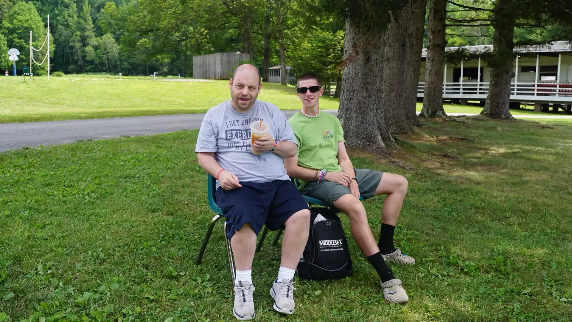 Two men sitting on chairs outdoors. One holds food, other wears sunglasses. Green grass, trees.