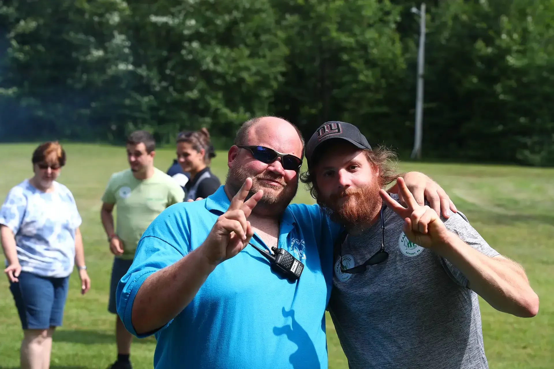 Two men smiling, flashing peace signs, embraced outdoors. People and trees in the background.