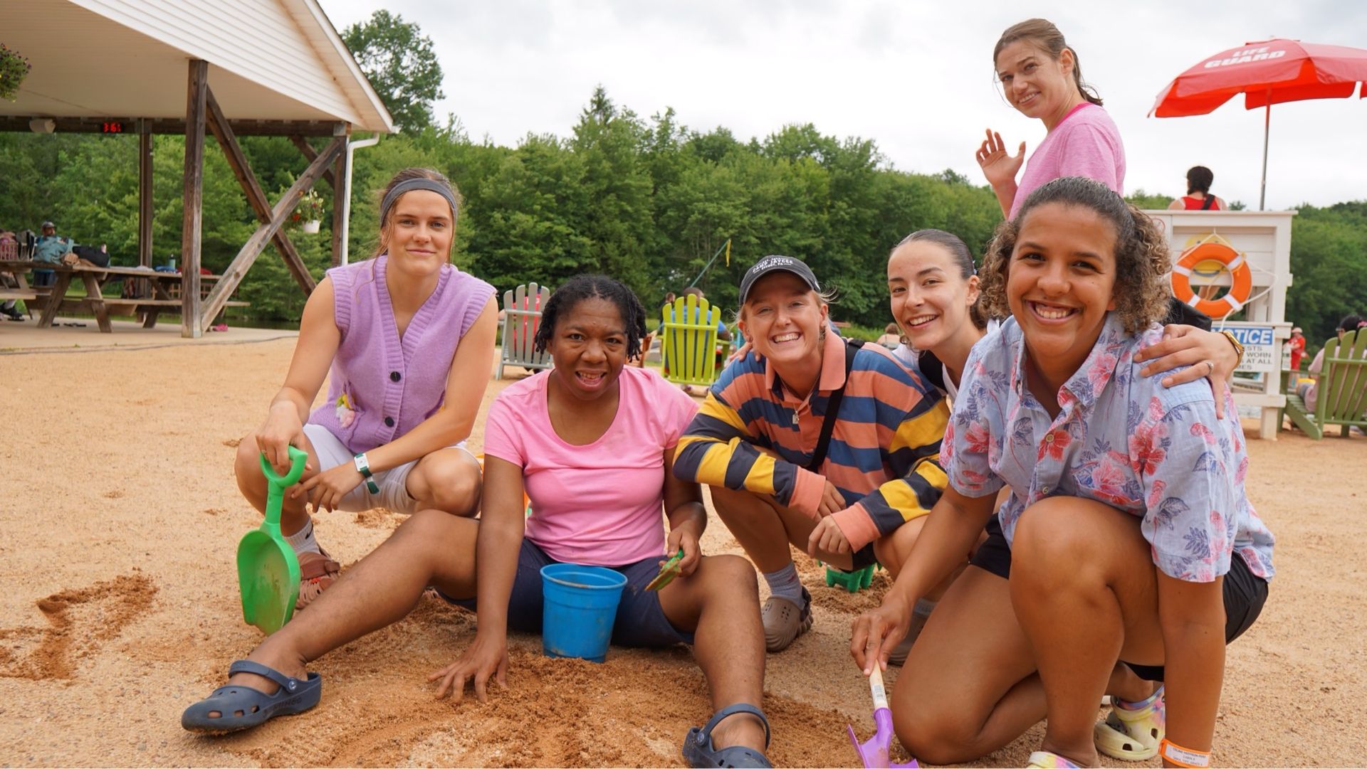 Group of smiling people in sand, some holding shovels and buckets, under a pavilion, trees in the background.