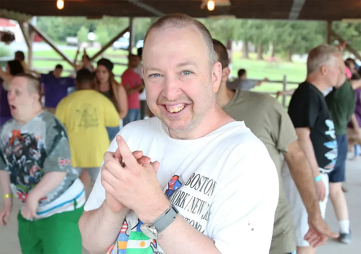 Man with a white shirt smiles, clapping. People dance in the background, under a pavilion.