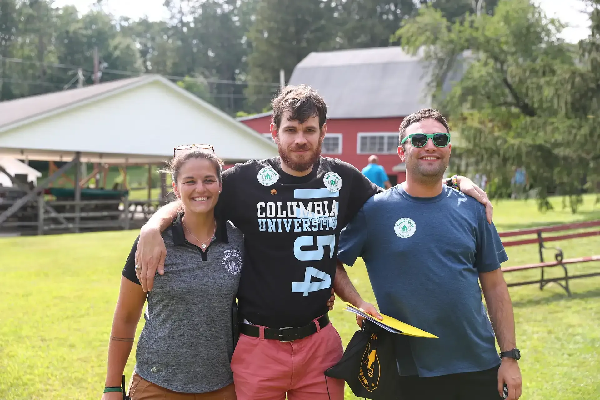 Three people with arms around each other posing outside. One wears a Columbia University shirt. Green field, buildings in background.
