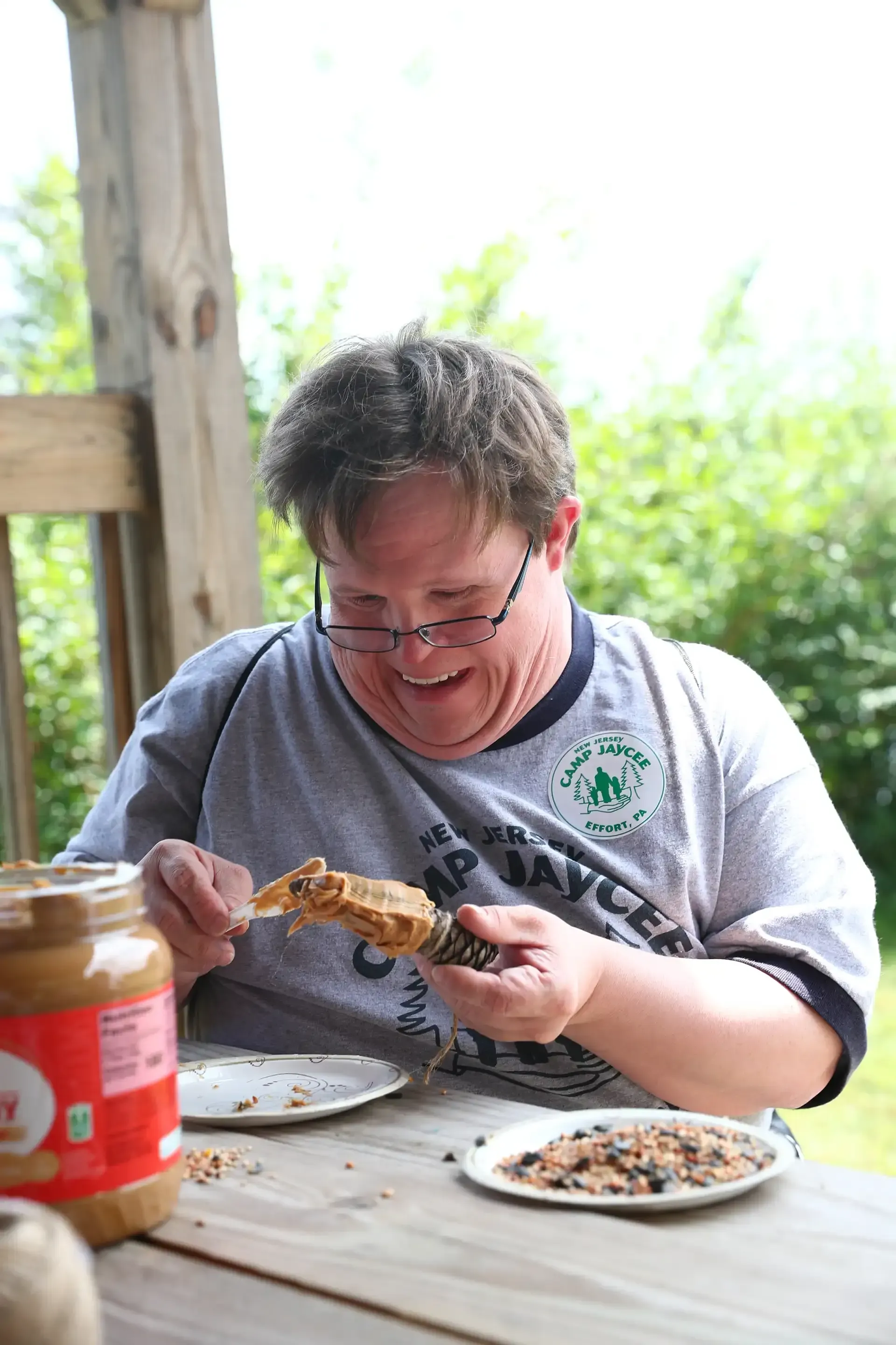 Man smiles while spreading peanut butter on a bird feeder at a wooden table outdoors.