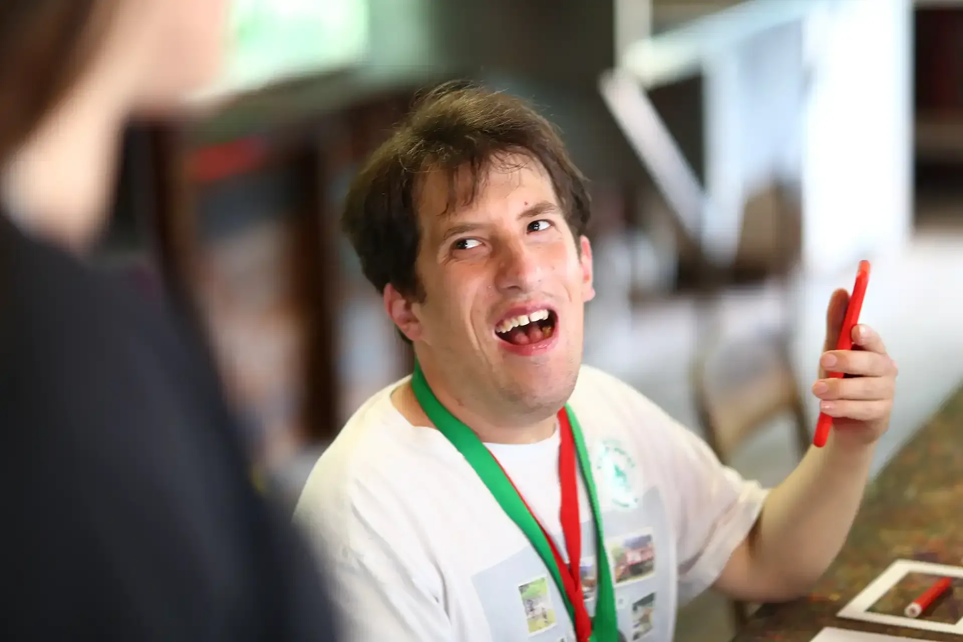 Man with Down syndrome smiling, holding a red stylus, wearing a lanyard, and looking towards an unseen person.