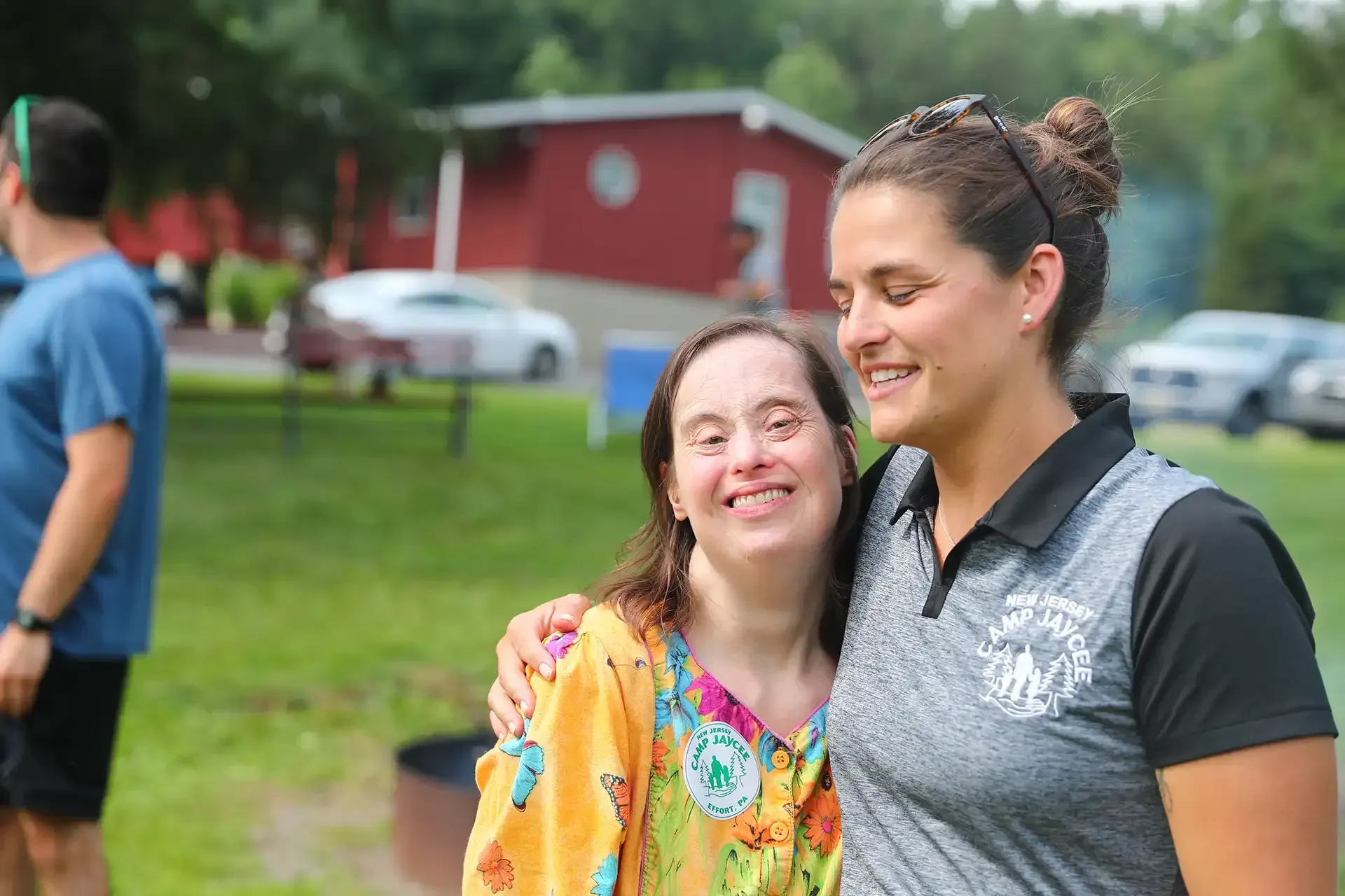 Woman with Down syndrome smiles, arm around another woman's shoulder outdoors. Cars and a red building are in the background.