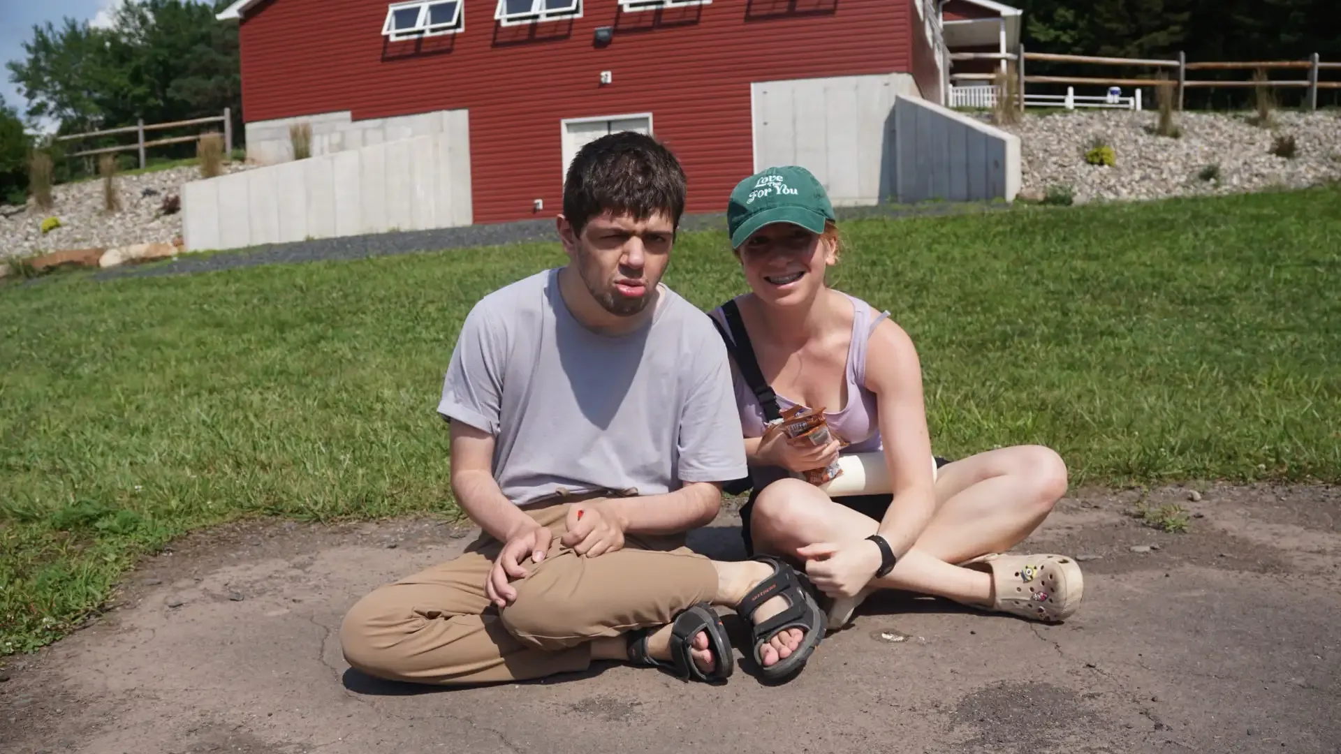 Two people sitting cross-legged on pavement in front of a red building and grassy area.