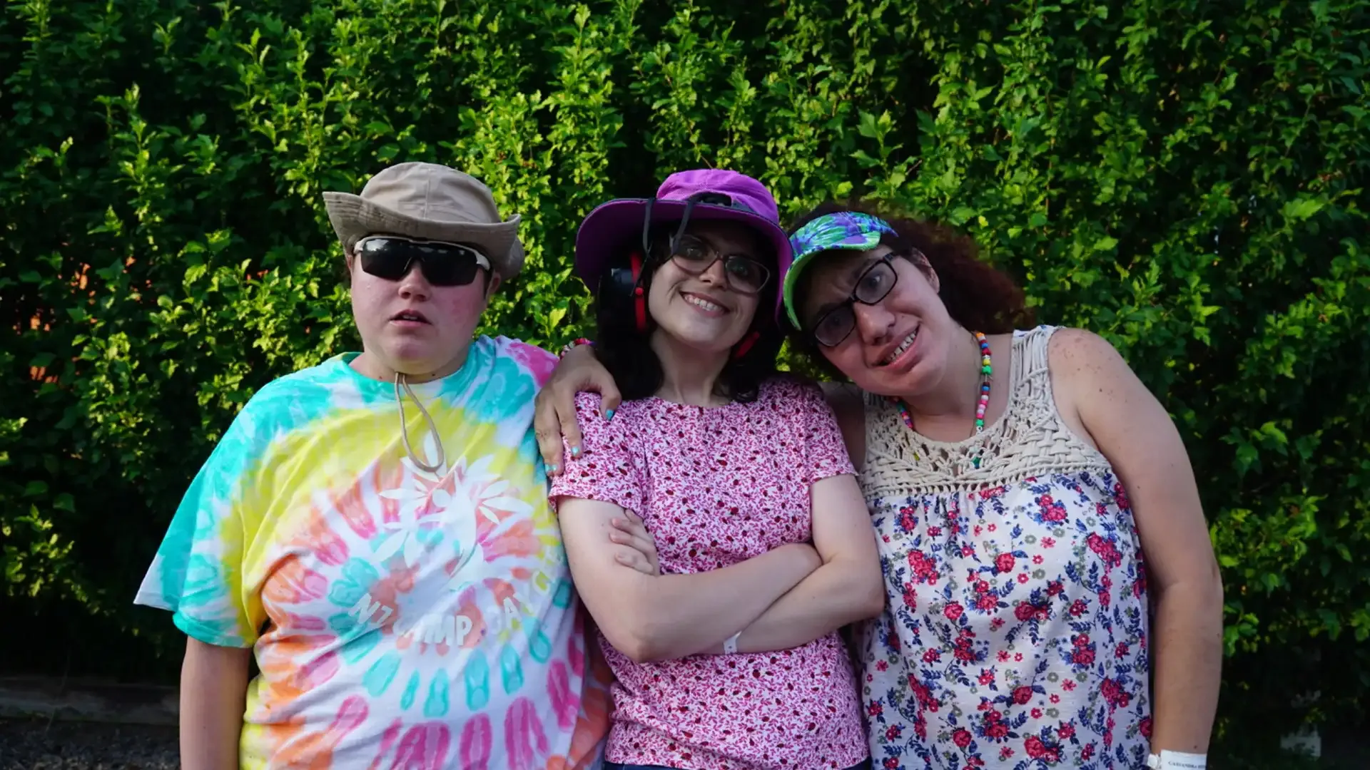 Three people smiling, wearing hats and sunglasses, posed in front of green foliage.