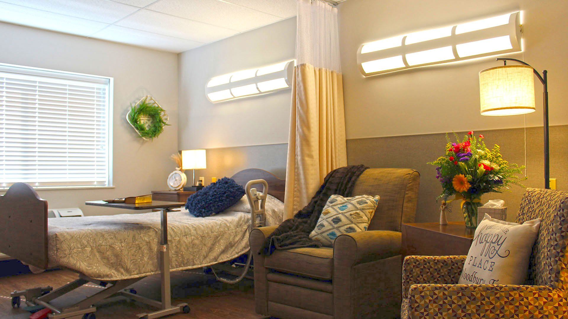 Hospital room with bed, armchair, flowers, and window.