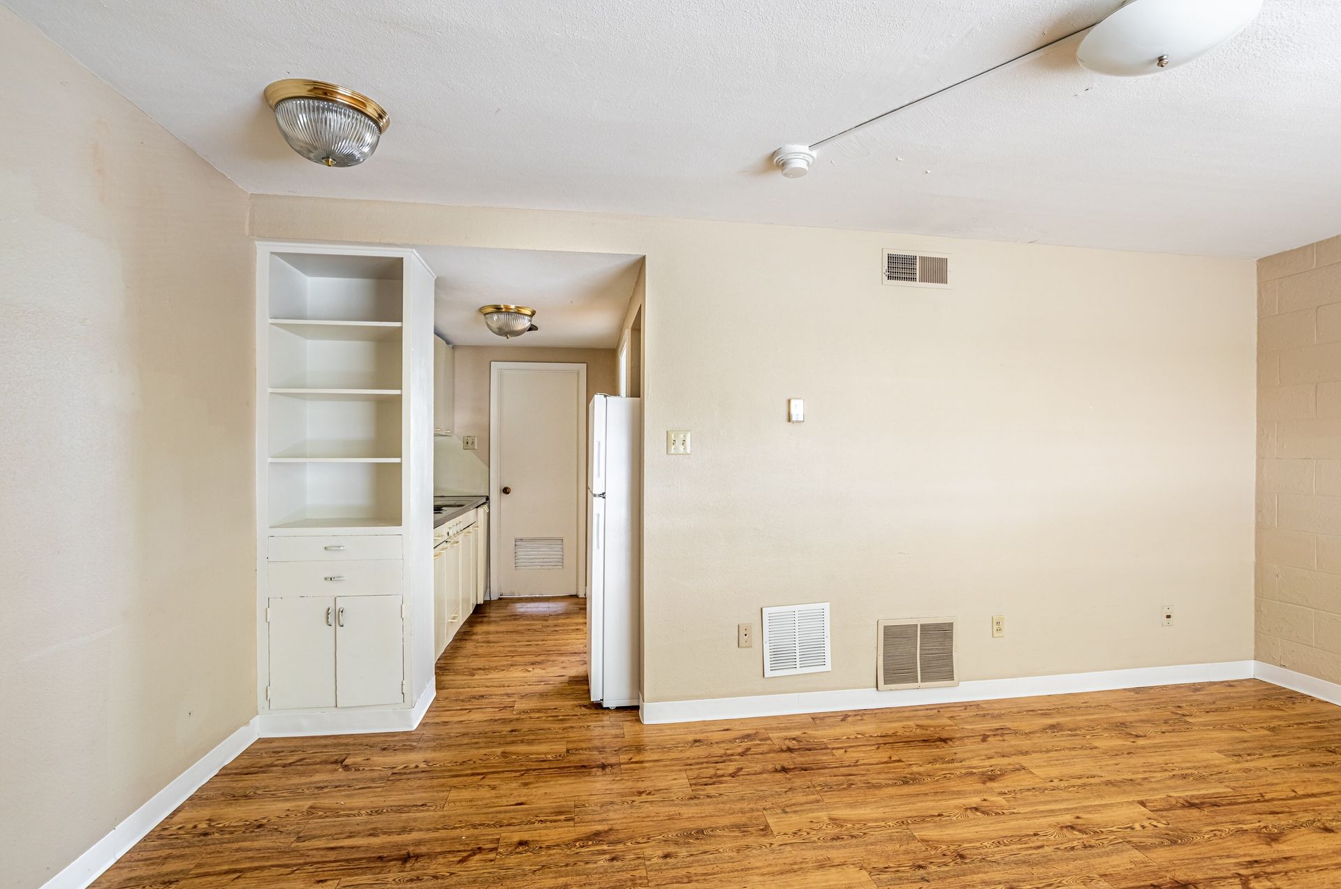 Empty apartment interior with kitchen, cabinets, and wood-look flooring. Beige walls and white appliances.