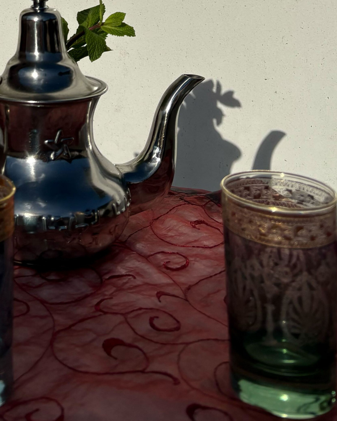 Théière en argent, verres à motifs et brin de feuillage sur un tissu rouge ; ombre sur un mur blanc.