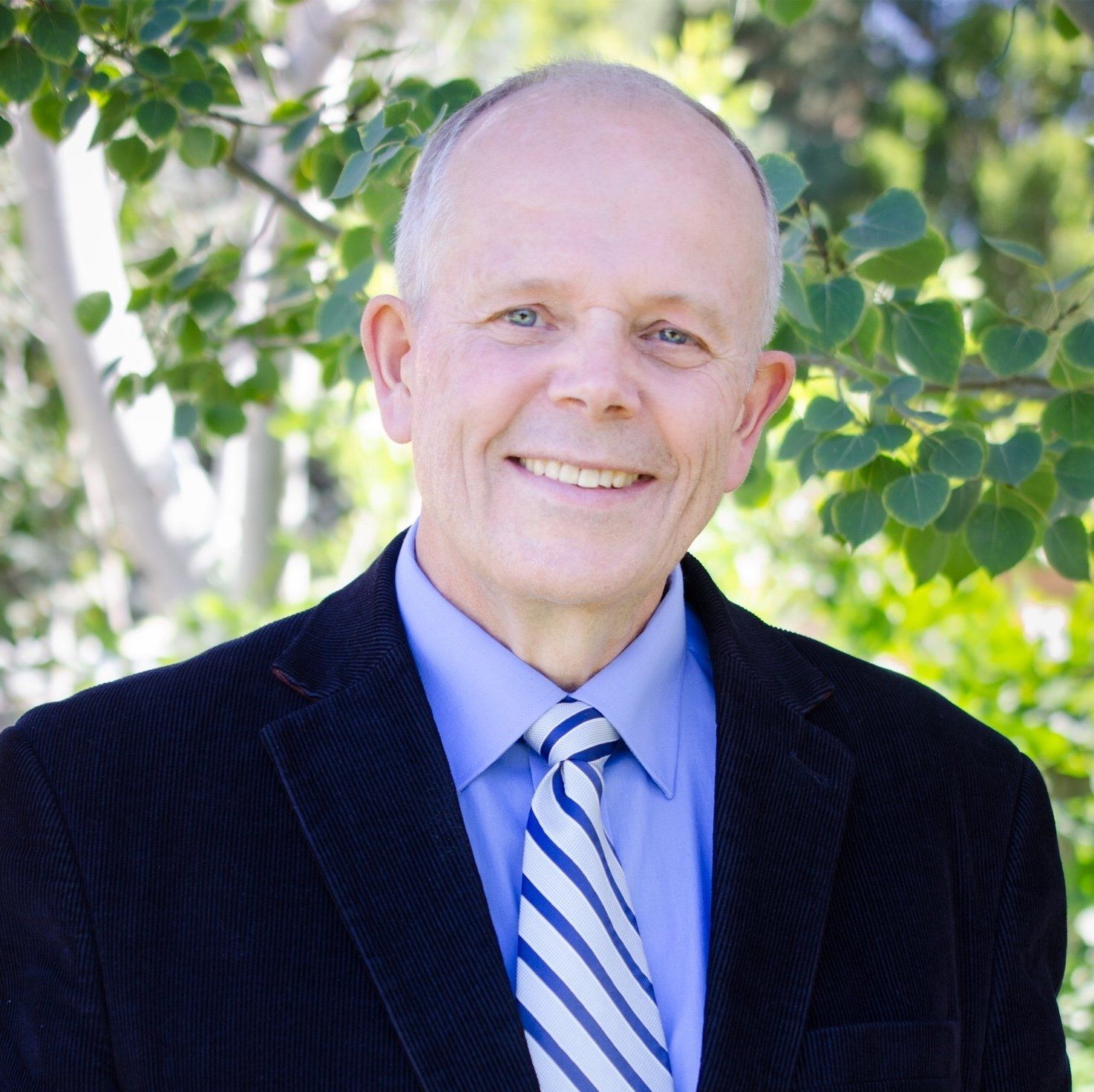 Smiling bald man in a navy suit and striped tie outdoors with leafy green background