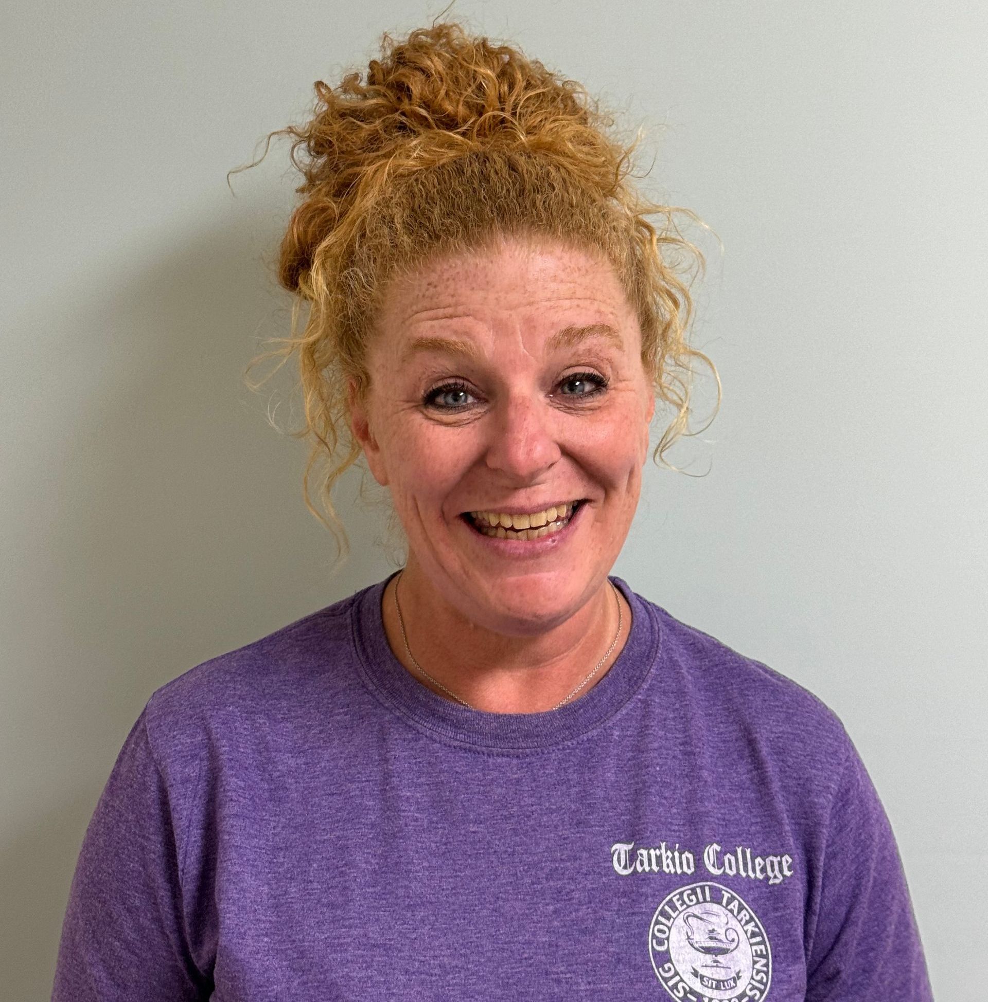 Smiling woman with curly red hair in a purple T-shirt against a plain gray wall