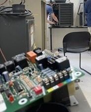 Close-up of a circuit board on a table, with lab equipment and a chair in the background.
