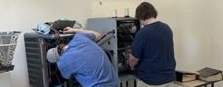 Two people working on open computer cases at a desk in a workshop.