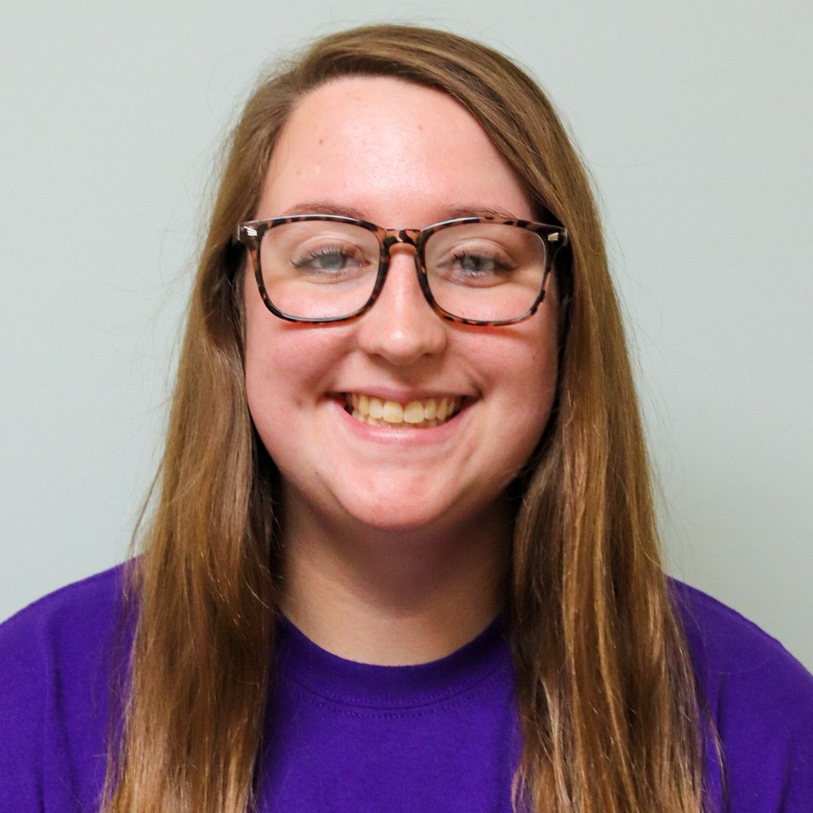 Smiling person wearing glasses and a purple shirt against a plain light background