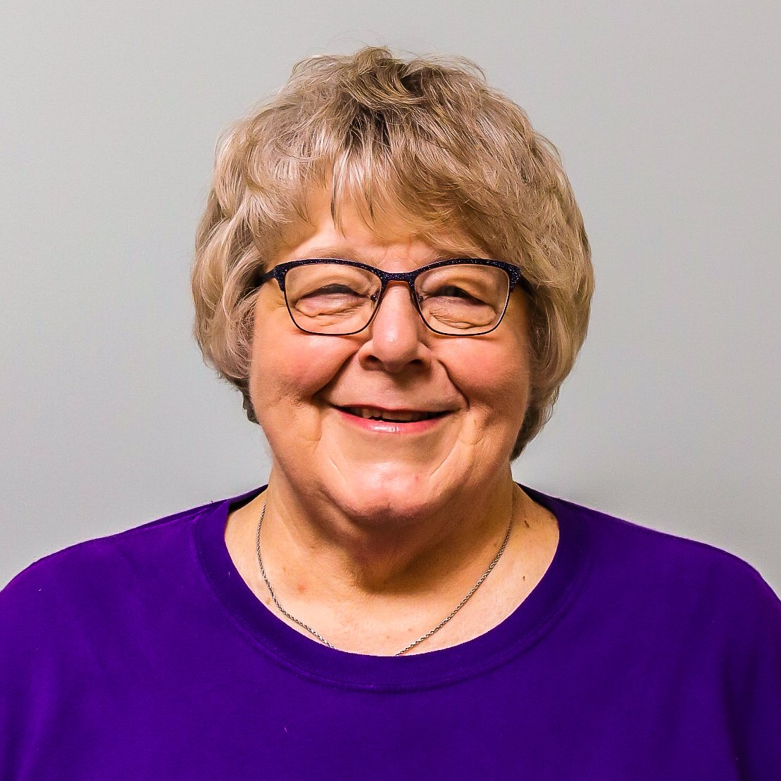 Smiling person with short blond hair and glasses, wearing a purple shirt against a light gray background
