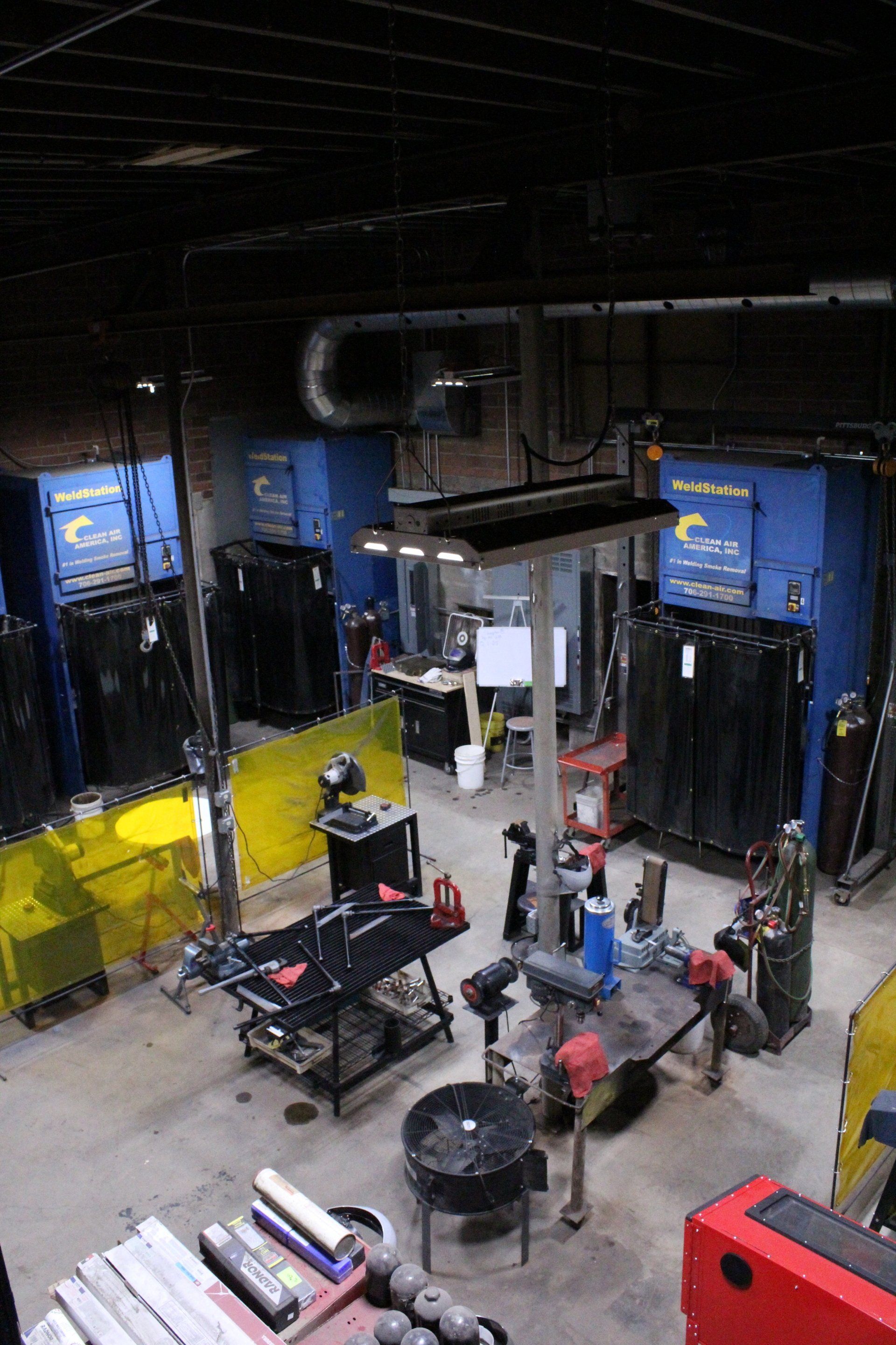 Indoor industrial workshop with blue machines, yellow barriers, and people working around equipment.