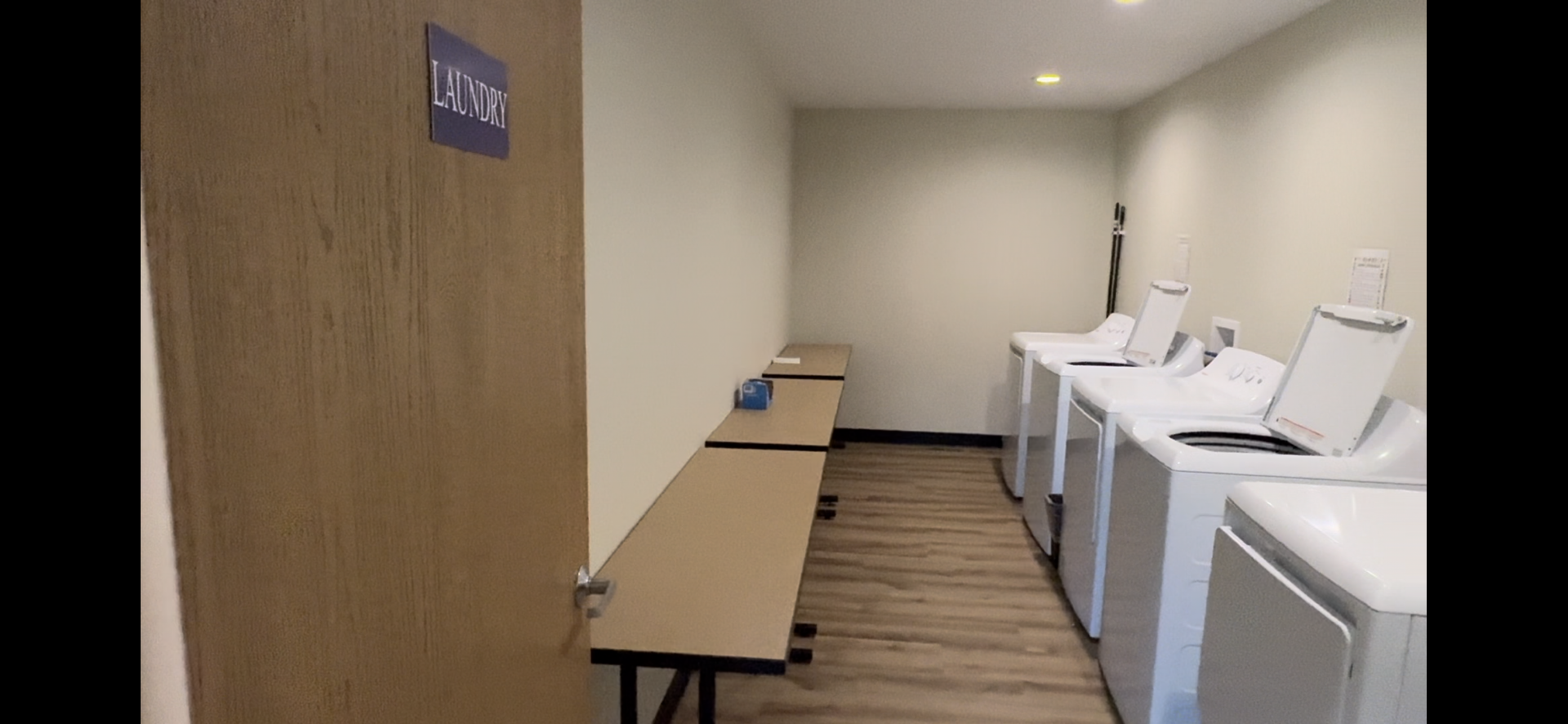 Laundry room with white washers, folding tables, and wooden door in a narrow hallway.