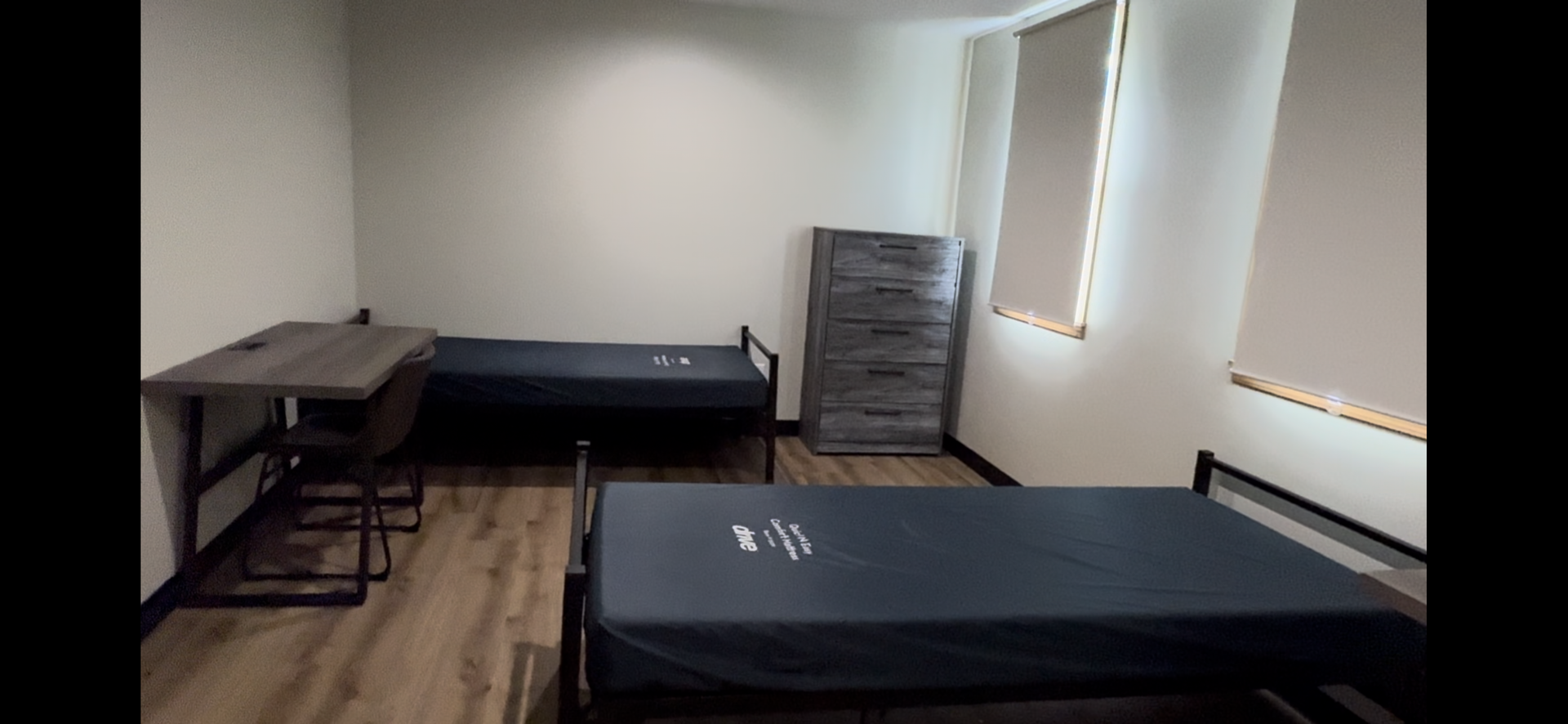 Sparse room with two beds, a desk, a dresser, and sunlight from windows