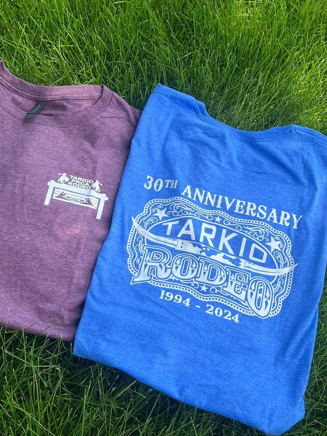 Two folded graphic T-shirts on grass, one purple and one blue with anniversary logos and text.