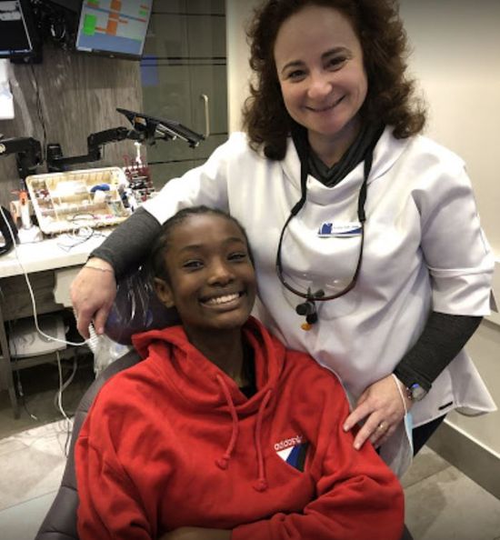 Woman in red hoodie smiles in a dental chair, another woman in white coat smiles behind her in a dental office.