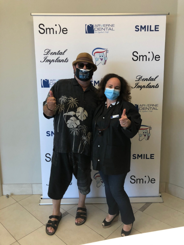 Man and woman give thumbs up in front of a banner. Man wears hat and sandals, woman in a mask.