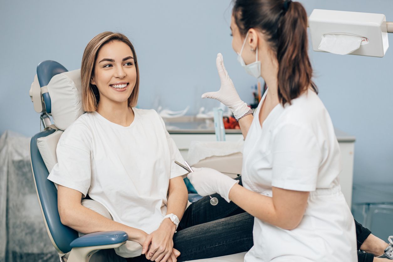 Woman smiling in dental chair, conversing with dentist wearing gloves and mask. Blue walls, dental equipment.